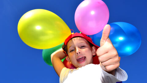 Kind steht mit bunten Luftballons vor dem blauen Himmel und streckt den Daumen nach oben