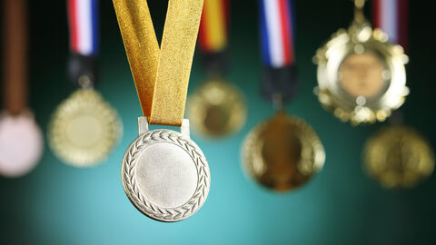 Silbermedaille hängt an einem goldenen Band im Vordergrund. Im Hintergrund sieht man verschwommen mehrere Medaillen hängen.