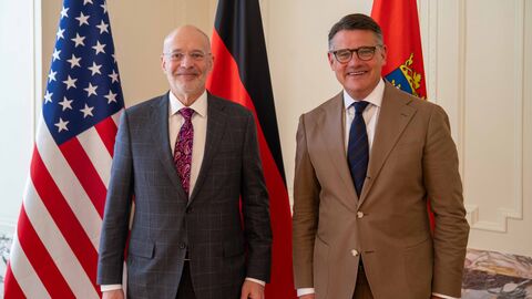 Zwei Männer stehen vor Flaggen der USA, Deutschland und Hessen.