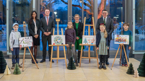 Vier Kinder stehen neben Bildern auf Staffeleien. Dahinter stehen 4 Erwachsene. 