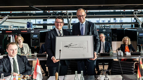 Ministerpräsident Rhein und Bundeskanzler Merz stehen nebeneinander und halten ein gerahmtes Bild.